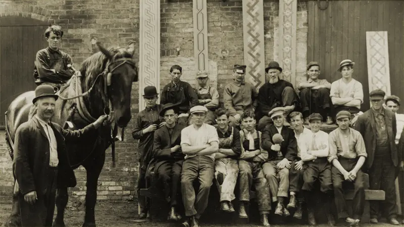 Horse stands with group of men against brick wall.