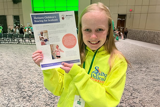 A patient stands in a large event hall holding a Shriners Children’s brochure about scoliosis bracing. The patient wears a bright yellow hoodie and displays the brochure toward the camera, with rows of chairs and event attendees visible in the background. The brochure includes photos and information about scoliosis treatment, highlighting the patient's connection to orthopedic care and advocacy.
