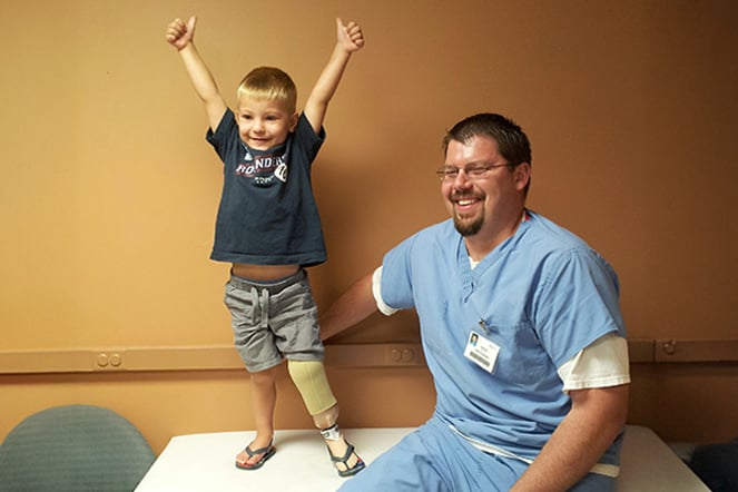 a happy child wearing a leg prosthetic giving two thumbs up next to a man in hospital scrubs