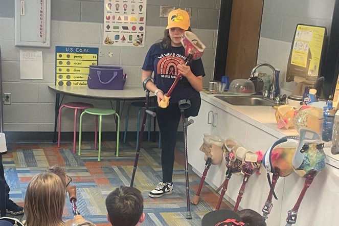 a woman holding up a prosthetic leg in front of a group of children in an elementary classroom