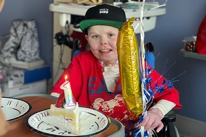 a child with burn scars smiling in front of a piece of cake with a candle and balloon shaped like the number 1