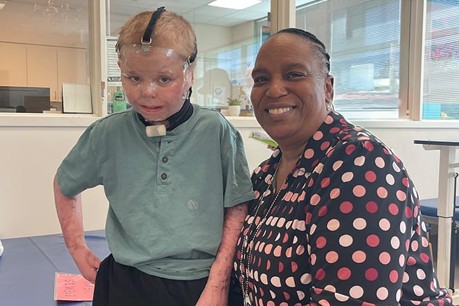 a child wearing a burn mask with an adult woman kneeling beside him