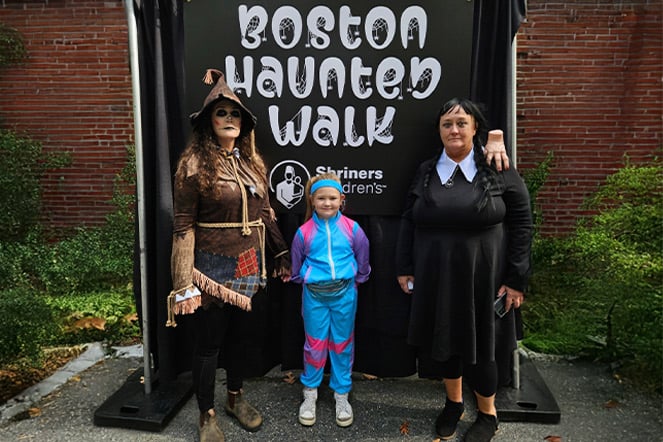 three people in Halloween costumes in front of a sign that reads "Boston Haunted Walk"