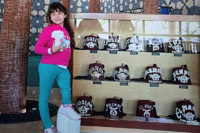 a child wearing a shoe with a custom lift stands next to a glass display of fezzes