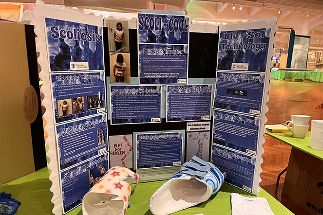 Tri-fold presentation board titled "Scoli‑Cool" and "Blu‑Sol Technology" explaining scoliosis and brace innovation, with photos, diagrams, and two prototype scoliosis braces displayed on a table in an exhibition space.