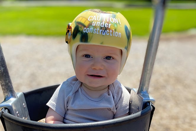 an infant sitting in a swing wearing a cranial molding helmet that reads "Caution: Under Construction" 