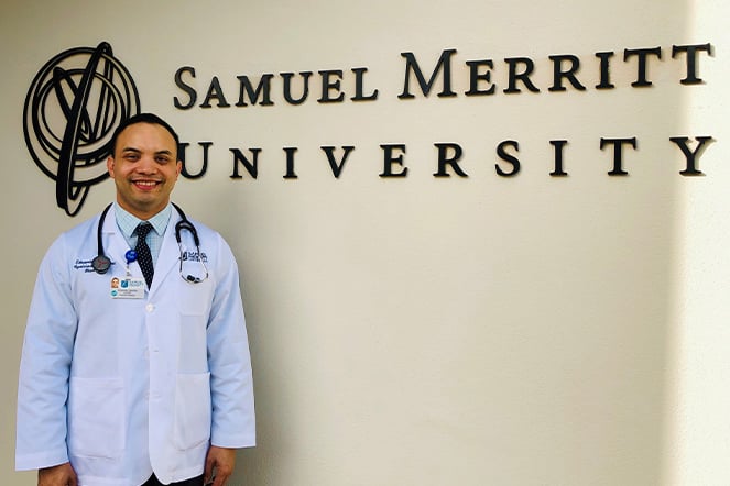 a man in a lab coat smiling in front of a sign that reads "Samuel Merritt University"
