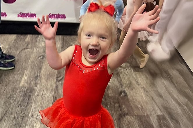 a toddler in a tutu raising their arms in a busy room