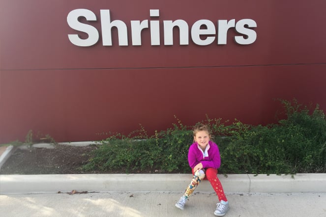 a young child with a prosthetic leg featuring designs of the character Wonder Woman sitting underneath a sign that reads "Shriners"