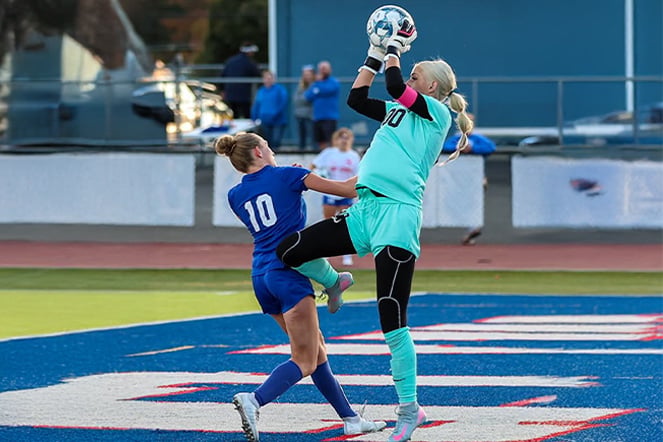 a goalie jumps and catches a soccer ball in front of an opponent