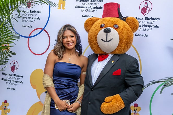 Person in a navy blue formal outfit stands beside Fezzy Bear, the Shriners Hospitals for Children mascot, in front of a branded step-and-repeat backdrop. Fezzy Bear wears a black suit jacket, red bow tie, red pocket square, and a red fez hat. The backdrop features bilingual Shriners Hospitals for Children Canada logos and colorful circular graphics, while decorative palm leaves frame the photo on both sides.