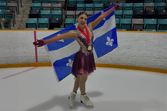Figure skater standing on an indoor ice rink with arms outstretched while holding a large blue-and-white Quebec flag behind their back.