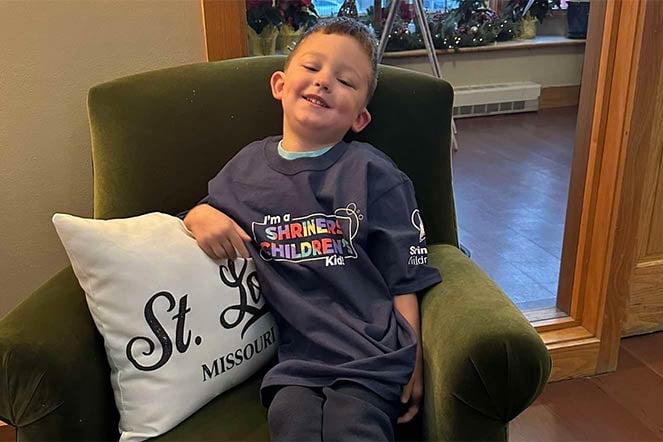 orthopedic patient lounges at home, leaning on pillow that has "St. Louis Missouri on it, T-shirt reads I'm a Shriners Children's Kid