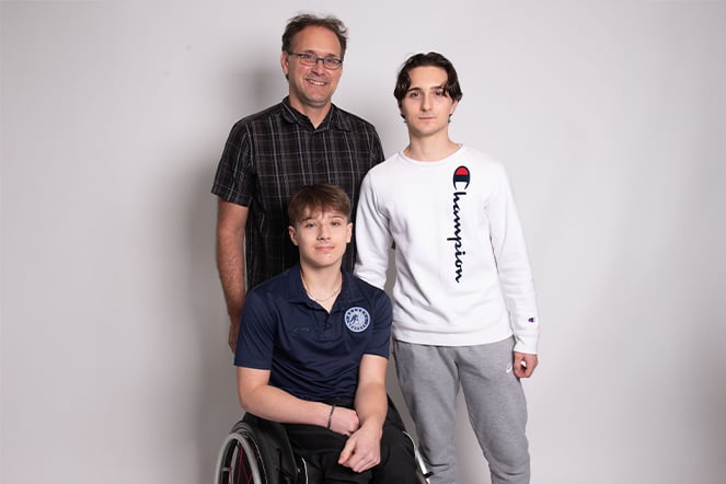 a portrait of a man standing next to a standing teenager and a teenager seated in a wheelchair