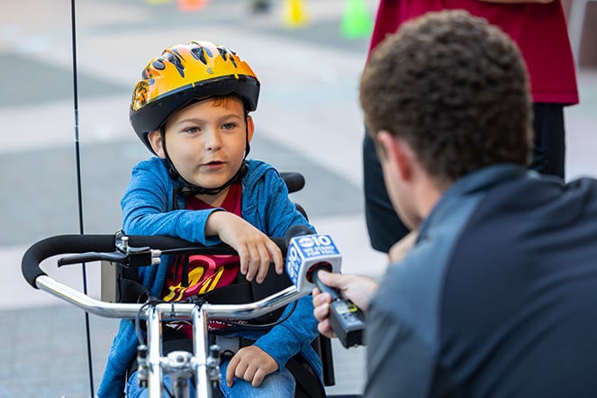 a child on an adaptive bicycle speaks into a microphone being held by a man