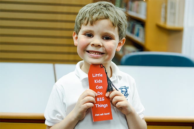 Young child standing in a library or classroom setting holds a red ribbon-shaped bookmark with a tassel. The bookmark displays the text, “Little Kids Can Do BIG Things” and “Just ask David!”