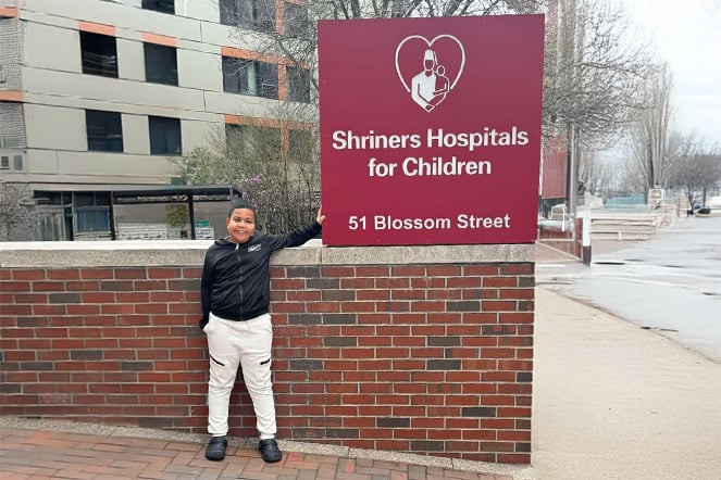 Child standing beside a large sign for Shriners Hospitals for Children outside the hospital campus. The burgundy sign displays the Shriners Hospitals for Children logo and the address, “51 Blossom Street.”