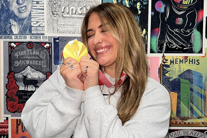 Person wearing a light gray sweatshirt poses indoors while holding a gold medal hanging from a red-and-white ribbon. The medal is displayed prominently near the person's face, and several rings are visible on the person's fingers. The background features a colorful wall covered with framed concert and event posters, including posters for Portland, Milwaukee, Memphis, and Trail Blazers-themed artwork, creating a vibrant, collage-style backdrop.