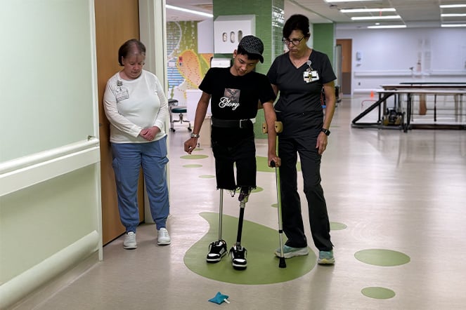 a child with two prosthetic legs walking with the assistance of one woman in scrubs while another woman looks on