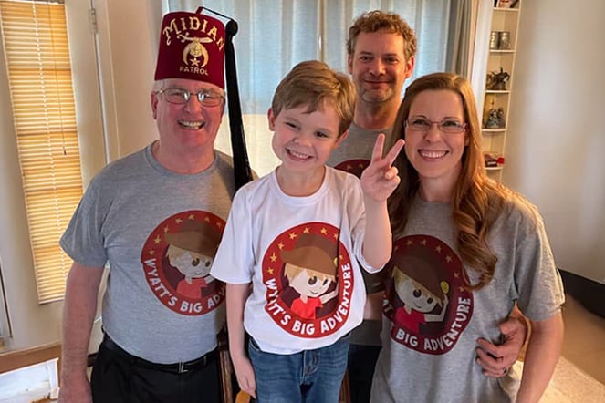 Four people stand together indoors wearing matching gray T-shirts featuring the Wyatt’s Big Adventure logo. One individual wears a maroon Shriners fez with gold embroidery reading “Midian Patrol.”