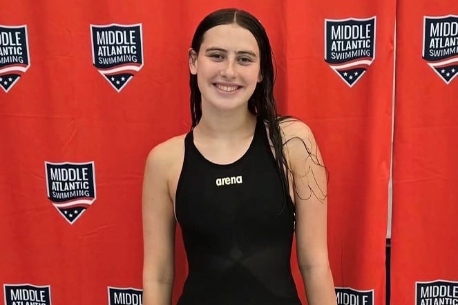Person in a black competitive swimsuit stands in front of a red backdrop featuring repeated Middle Atlantic Swimming logos. The athlete has wet hair and is posing for a photo at a swimming event. The backdrop displays the text “MIDDLE ATLANTIC SWIMMING,” and the swimsuit features the Arena brand logo on the front.
