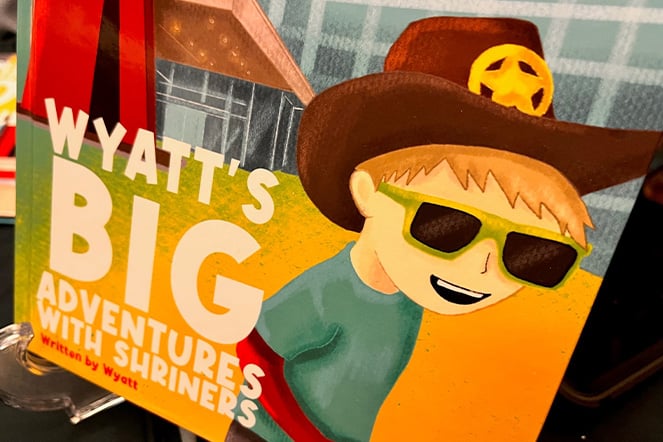Close-up of the cover of a children's book titled Wyatt’s Big Adventures with Shriners. The illustrated cover features a cartoon child wearing a large brown hat with a yellow emblem and green sunglasses, set against a colorful outdoor background with a building and bright yellow ground. The book title appears in large white letters, with the text “Written by Wyatt” near the bottom of the cover.