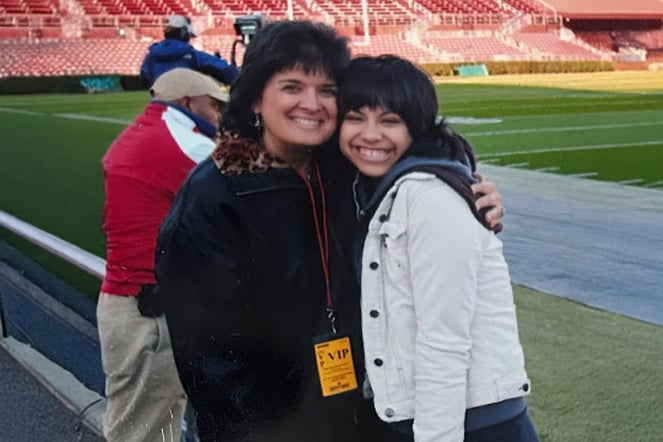 Two people pose together on the sideline of a football stadium, standing near the field with rows of red seats visible in the background. One person wears a VIP credential on a lanyard, and both are embracing for a photo during a stadium event.