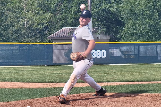 Baseball player in a gray jersey and white pants pitching from the mound during a game. The ball is visible above the player's throwing arm as the pitching motion is underway. A grass outfield, blue outfield fence with a “380” distance marker, trees, and a small building are visible in the background.