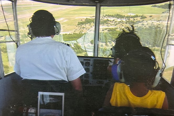A view from inside an aircraft cockpit shows three people wearing headsets seated at the controls and passenger seats while flying over a landscape below. Through the large front windows, fields, roads, and clusters of trees are visible, highlighting the aerial perspective during the flight.