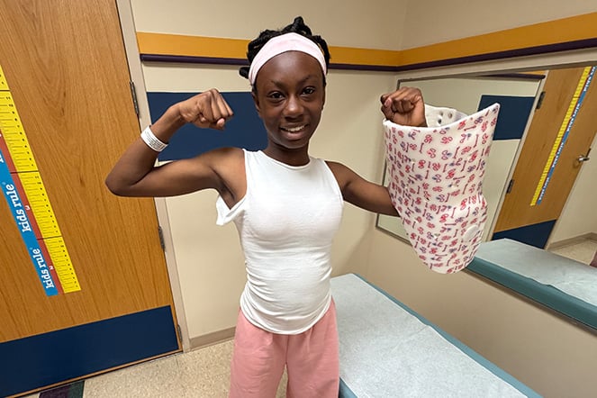 A patient stands in an exam room holding a scoliosis brace decorated with colorful patterns while flexing both arms.