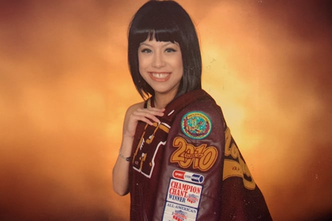 A person poses for a portrait while draped in a maroon letterman-style jacket featuring patches, awards, and embroidered lettering.