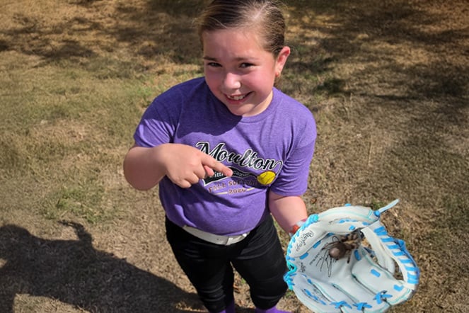 A youth softball player stands outdoors on a grassy area holding an open softball glove containing a large tarantula. The player wears a purple Morton Little League softball shirt, black softball pants and a white belt, while pointing toward the spider.