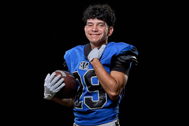 A football player in a blue uniform poses against a black background while holding a football. The player wears shoulder pads, gloves, and a jersey with the number 19, standing in a studio-style portrait that highlights the athletic gear and football.
