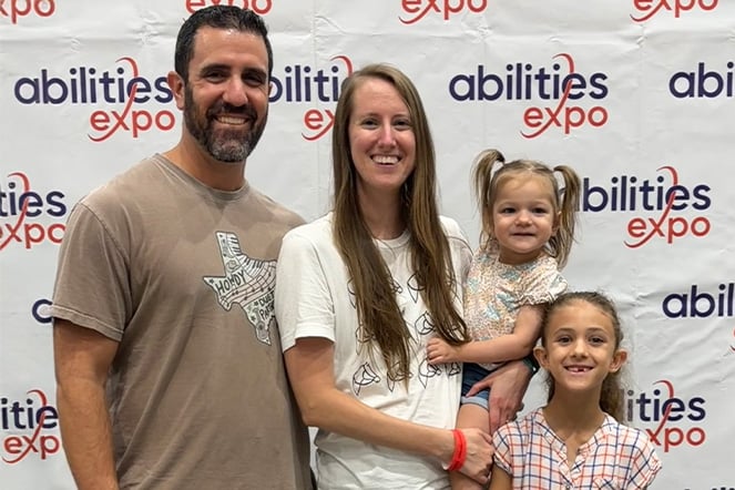 A group of four people pose in front of a step-and-repeat banner displaying the “Abilities Expo” logo. Two adults stand beside two children, with one adult holding a toddler.