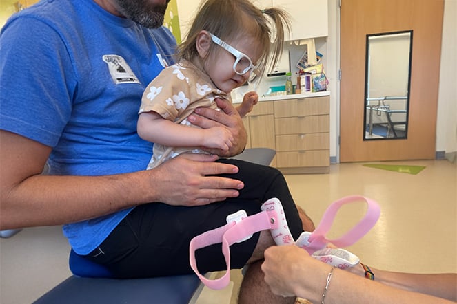 A child wearing white glasses sits on an adult’s lap in a clinical setting while a healthcare provider adjusts a pink orthopedic brace on the child’s lower leg and foot.
