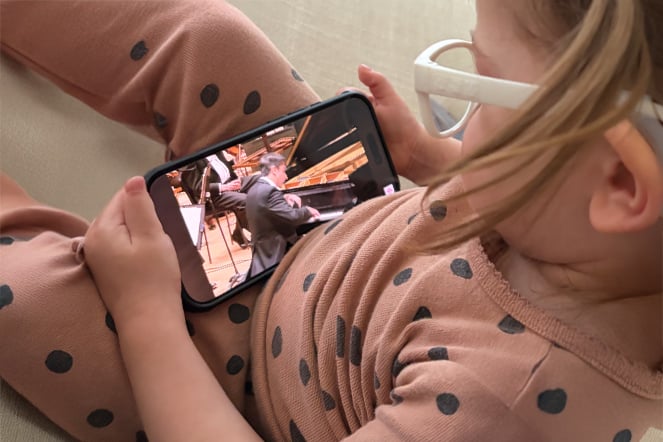 A child wearing white glasses and a tan outfit with dark polka dots reclines while holding a smartphone horizontally. The phone screen displays a pianist performing at a grand piano on a concert stage.