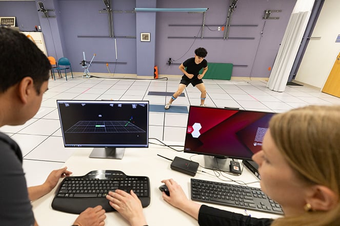Two clinicians sit at computer workstations in a motion analysis laboratory while observing a patient performing a movement exercise on a marked testing floor. Computer monitors display motion-tracking data and a 3D visualization, while specialized equipment and measurement markers around the room highlight the clinical gait and movement assessment setting.