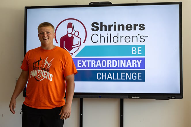 Person standing beside a large display screen showing the Shriners Children’s logo and the slogan “BE EXTRAORDINARY CHALLENGE.”