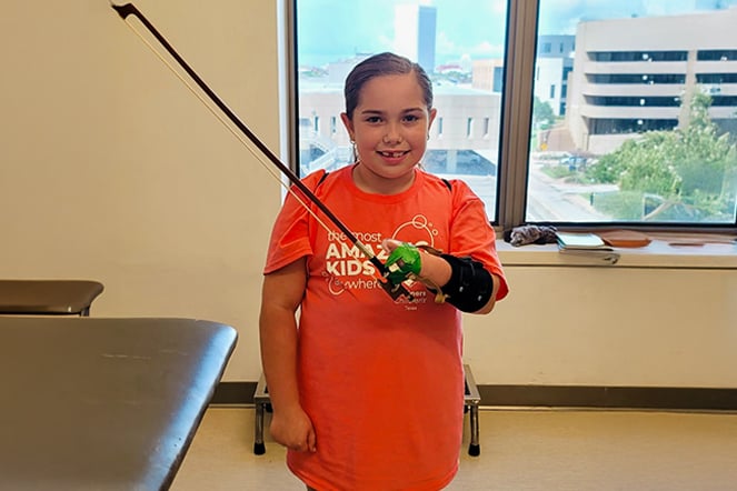 A child stands in a therapy room holding a violin and bow while wearing an orthotic support device on one arm and hand.