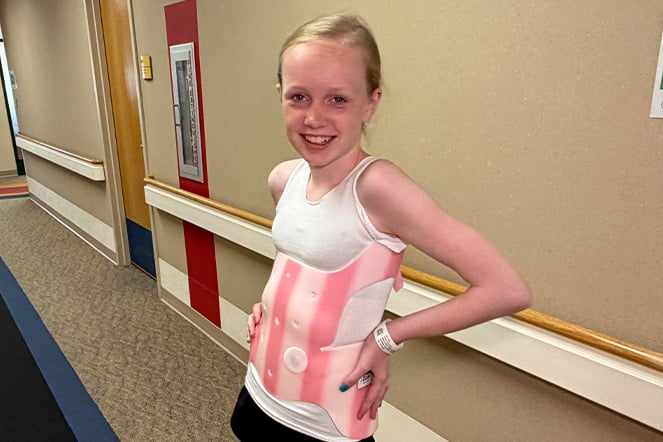 A patient stands in a hospital hallway wearing a pink-and-white scoliosis brace over a white tank top. The patient poses with hands on their hips, while colorful wall accents, handrails and a fire extinguisher cabinet line the corridor.