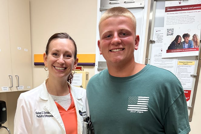 Two people stand side by side in a clinical office setting. One person wears a white medical coat over a coral shirt, and the other wears a green T-shirt with a flag graphic.