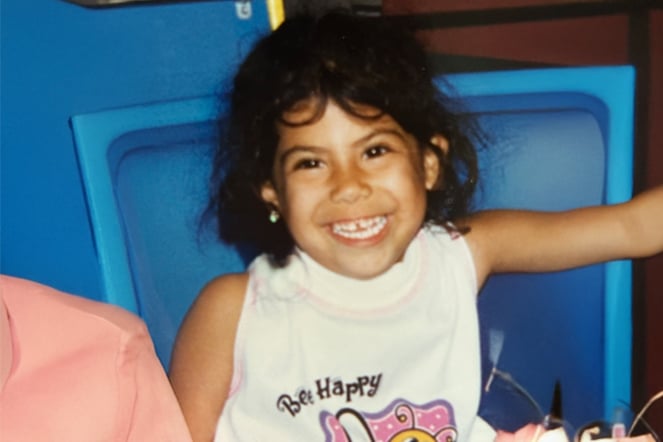 A young child sits in a blue plastic chair and smiles toward the camera.