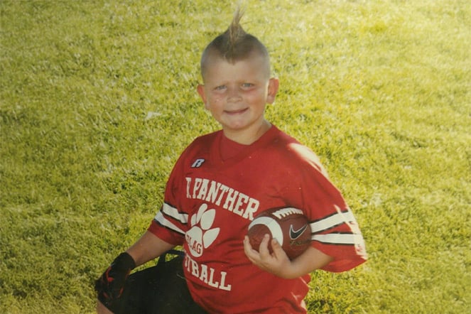 Young football player kneeling on a grassy field while holding a football and a helmet.