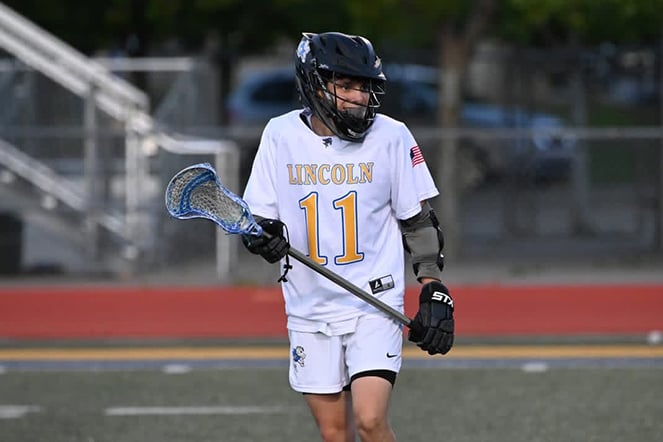 A youth lacrosse player wearing a white Lincoln jersey with the number 11 walks across a field while holding a lacrosse stick.