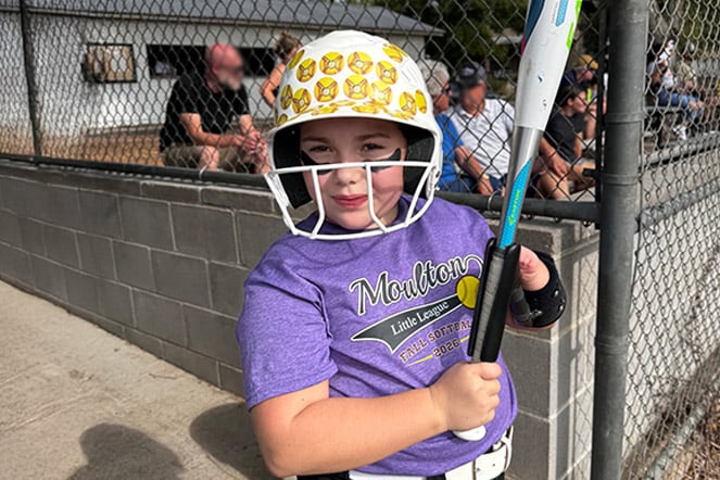A youth softball player stands near a chain-link fence holding a bat and wearing a white batting helmet decorated with yellow softball graphics. The player wears a purple Morton Little League softball shirt and protective batting gear, while spectators sit in the background watching activities at the ballfield.