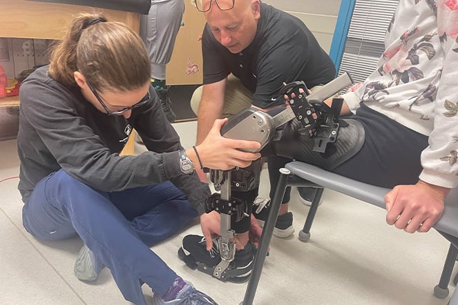 Two clinicians adjust and fit a specialized robotic-assisted leg brace on a seated patient during a therapy appointment. One clinician kneels beside the device while the other makes adjustments to the mechanical supports attached to the patient's leg and foot.