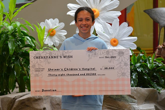 A patient stands indoors holding an oversized ceremonial donation check made out to Shriners Children’s Hospital for $38,000. The patient wears a light blue hoodie and stands in front of large decorative flower displays and lush greenery. The check, labeled “Cherryanne’s Wish,” is prominently featured in the foreground, highlighting a fundraising contribution to support pediatric healthcare.