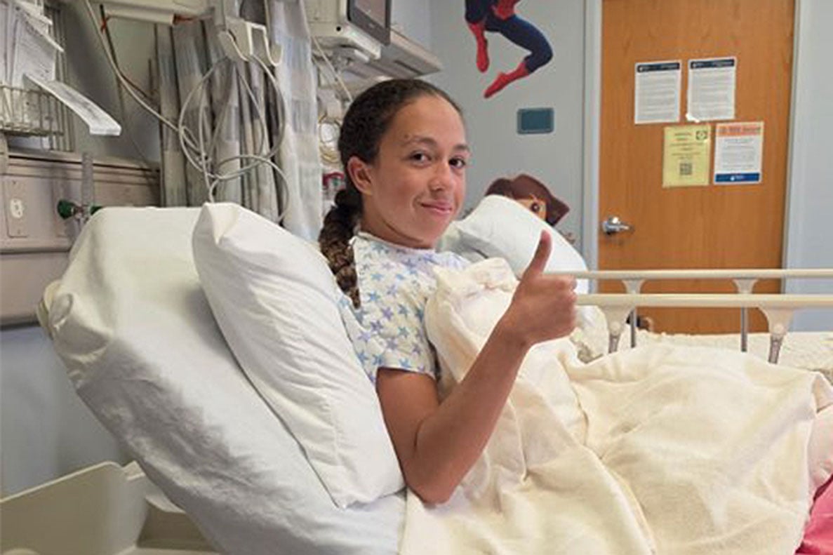 A patient sits upright in a hospital bed wearing a patterned hospital gown and gives a thumbs-up toward the camera. Medical equipment, monitors and cables are mounted along the wall beside the bed, while colorful character decals and informational notices are visible in the room.
