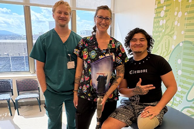 A patient wearing a lower-limb prosthesis poses with two prosthetics and orthotics team members in a hospital clinic. The patient sits on an examination table and wears a black T-shirt and patterned shorts, while the clinicians stand alongside in scrubs.