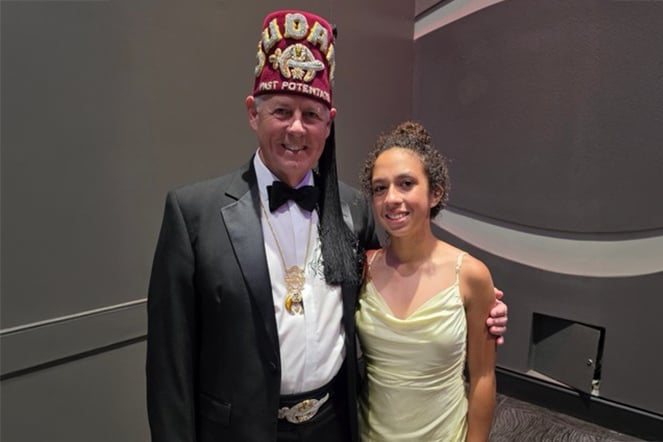 A patient and a Shriners representative pose together for a photo at a formal event. The Shriners representative wears a tuxedo, ceremonial fez and medallion, while the patient wears a light-colored dress.
